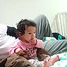 Taliah joined the competition — help win amazing prizes! baby, child, infant, sitting, bed, bedroom, blanket, pillow, clothing, pink_shirt, patterned_pants, bare_feet, curly_hair, face, hand, finger, looking_away, indoors, furniture, portrait