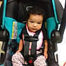 baby, child, toddler, stroller, car_seat, safety_harness, seated, portrait, indoor, serious, curly_hair, pink_clothing, black_clothing, turquoise, baby_transport, harness, face, hands, legs, photography