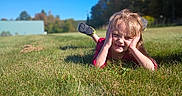 Via is registered to the contest to win money with this photo: child, grass, outdoor, blue_sky, playful, hands_on_face, casual_clothing, shoes, greenery, sunny, daylight, nature, portrait, young, person, field, expression, relaxed, fall, trees