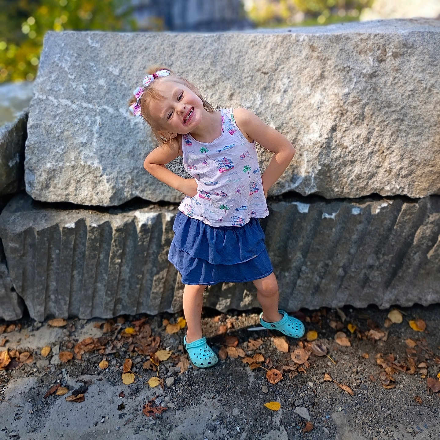 Aviana is registered to the contest to win money with this photo: child, girl, outdoor, rocks, smile, playful_pose, autumn_leaves, blue_skirt, tank_top, crocs, sunlight, nature, happy, standing, hair_bow, casual_clothing, portrait, young, daytime, background_blur