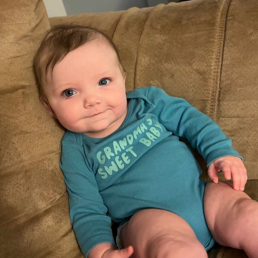 Rowyn is registered to the contest to win money with this photo: baby, blue_clothing, child, couch, cute, expression, face, furniture, hand, home, indoor, infant, leg, onesie, person, relaxing, seated, skin, smiling, young_child