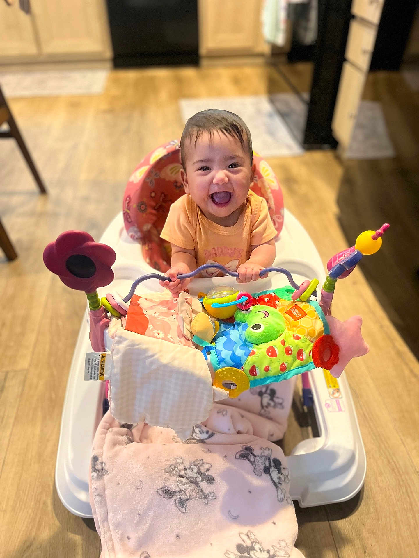 Alanna is registered to the contest to win money with this photo: baby, walker, toy, blanket, smiling, child, indoor, floor, wooden_floor, kitchen, happy, person, colorful, playtime, seat, infant, cute, clothing, face, furniture