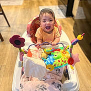 Alanna is registered to the contest to win money with this photo: baby, walker, toy, blanket, smiling, child, indoor, floor, wooden_floor, kitchen, happy, person, colorful, playtime, seat, infant, cute, clothing, face, furniture