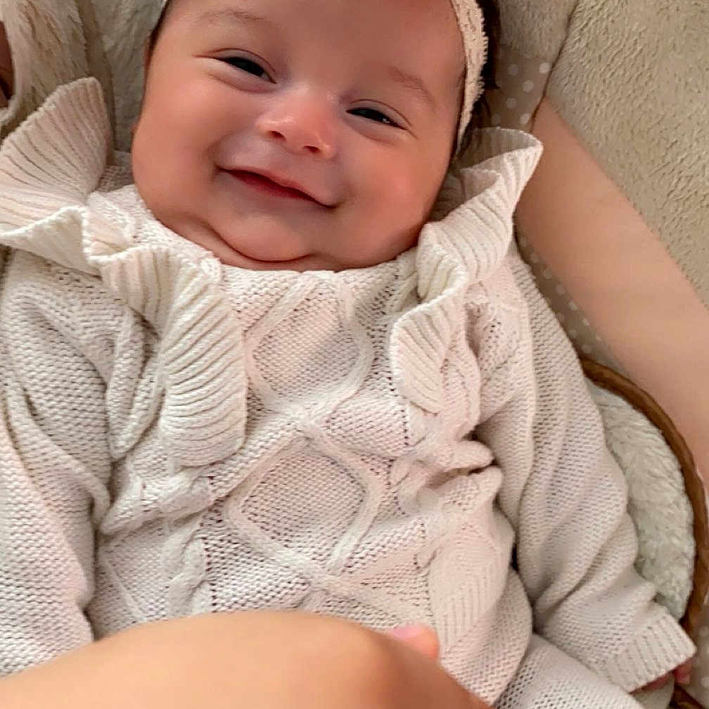 Madelyn joined the competition — help win amazing prizes! baby, child, closeup, comfort, cozy, cushion, cute, hand, happy, headband, indoors, infant, knitted_outfit, pink_flower, portrait, resting, ruffles, smiling, soft_texture, white_clothing