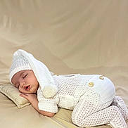 Marina a rejoint le concours — aidez-le/la à gagner de superbes lots ! baby, infant, newborn, sleeping, knit_hat, pom_pom, onesie, knitwear, pillow, beige_background, cozy, portrait, studio, soft_lighting, peaceful, hand, legs, face, clothing, lying_down