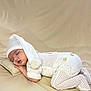 baby, infant, newborn, sleeping, knit_hat, pom_pom, onesie, knitwear, pillow, beige_background, cozy, portrait, studio, soft_lighting, peaceful, hand, legs, face, clothing, lying_down