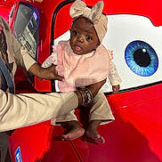 Ziv joined the competition — help win amazing prizes! baby, child, person, red_car, cartoon_eye, headband, pink_bib, clothing, barefoot, adult_hands, playful_expression, indoor, toy_car, reflection, bracelet, long_sleeves, pants, cute, portrait, holding