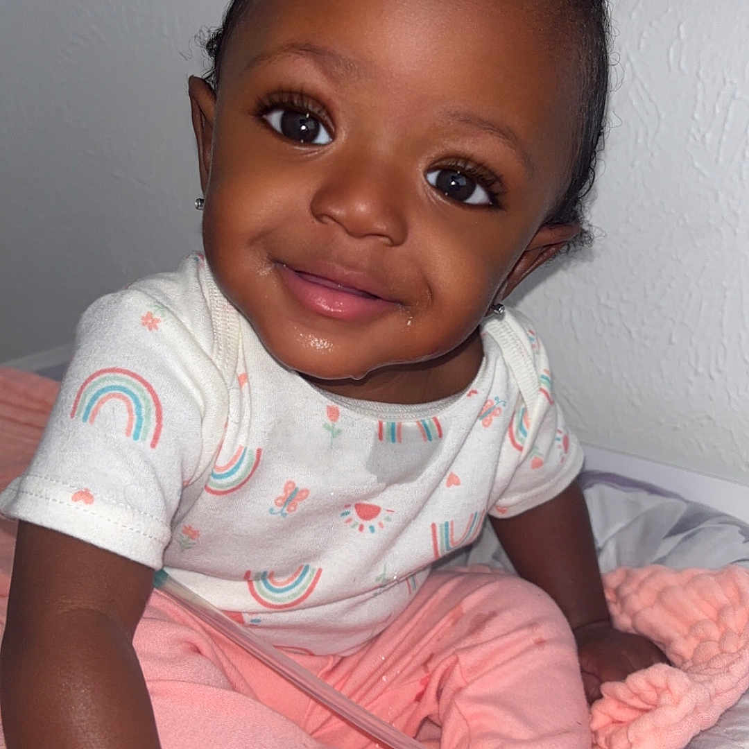 Ziv joined the competition — help win amazing prizes! baby, bed, blanket, child, clothing, curly_hair, cute, earrings, face, foot, hair_tie, hand, indoors, painting, pink_pants, portrait, rainbow_pattern, sitting, smiling, wall