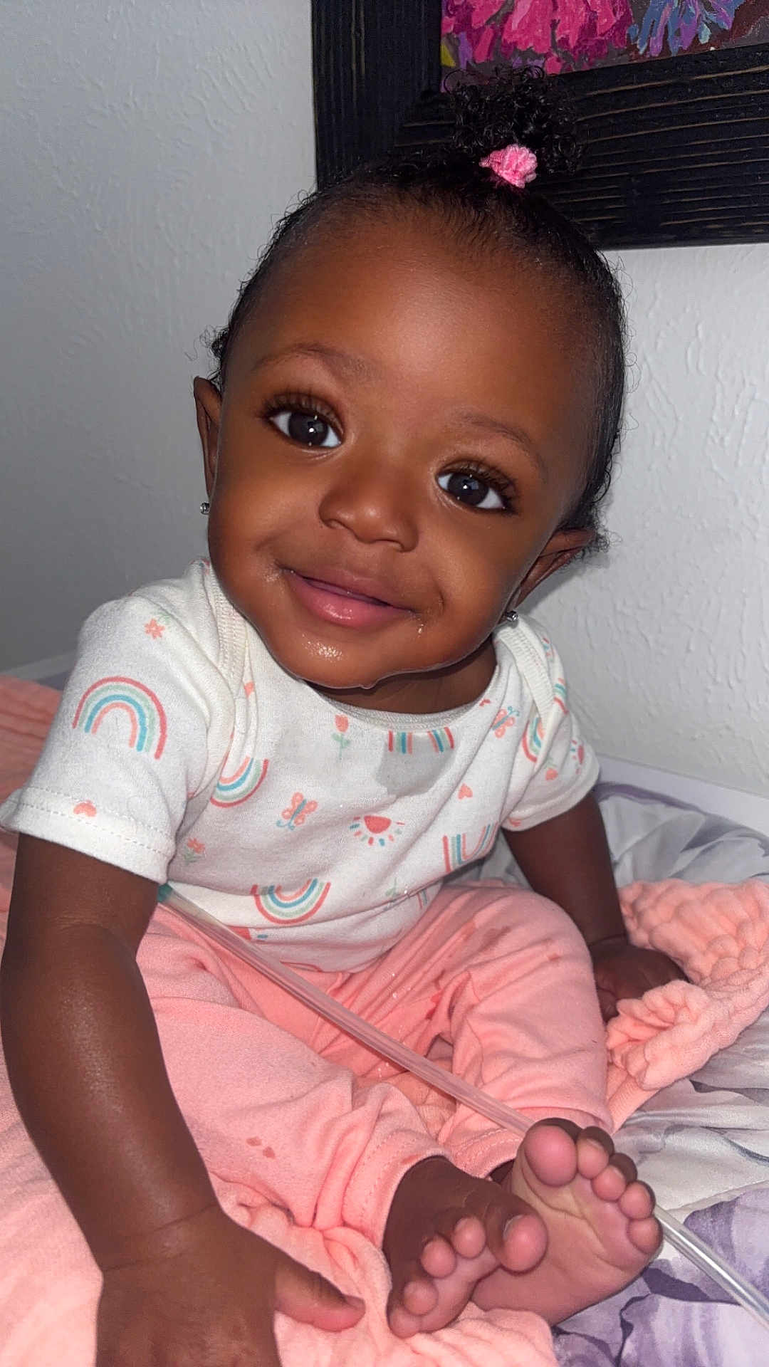 Ziv joined the competition — help win amazing prizes! baby, child, smiling, sitting, indoors, clothing, rainbow_pattern, pink_pants, blanket, bed, foot, hand, earrings, curly_hair, hair_tie, face, portrait, wall, painting, cute
