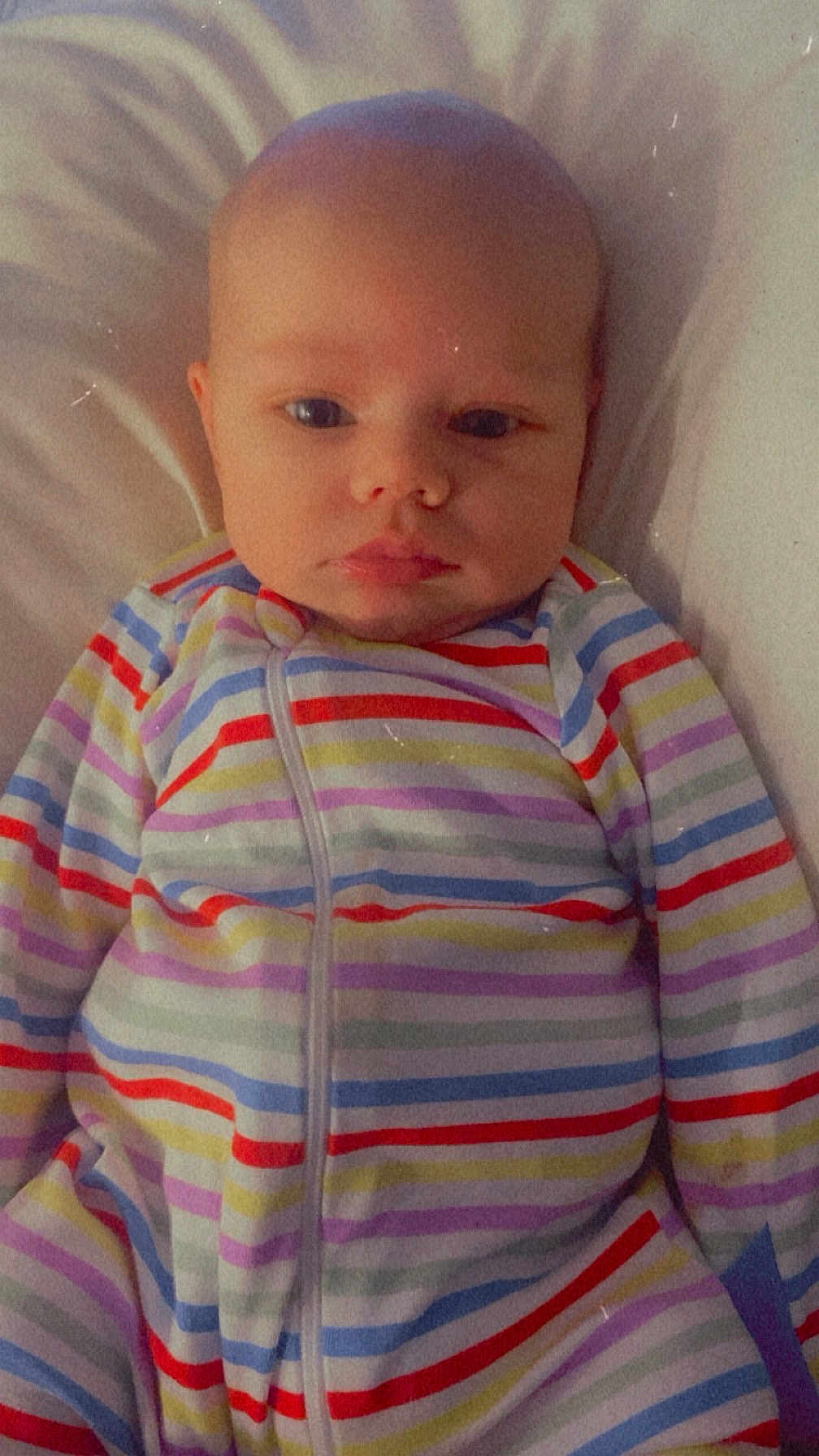 Oaklyn is registered to the contest to win money with this photo: baby, child, infant, face, striped_clothing, onesie, lying_down, portrait, serious_expression, soft_surface, person, young_child, sleepwear, colorful, cute, indoors, closeup, head, skin, calm