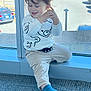 toddler, child, curly_hair, sitting, window, car, parking_lot, blue_sock, beige_pants, white_sweatshirt, bear_design, happy, indoor, daylight, casual_clothing, smiling, young_child, floor_carpet, real_photo, natural_light