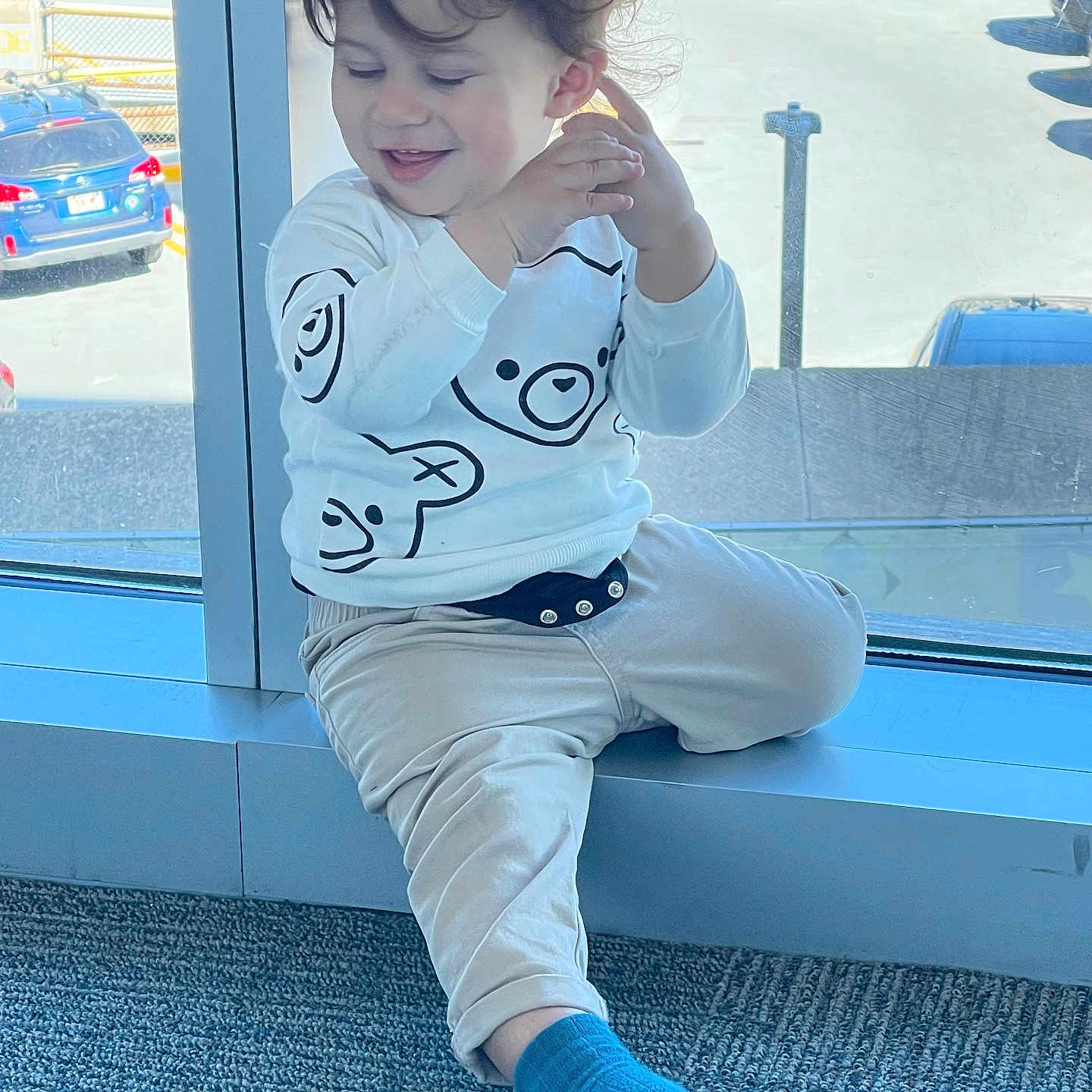 Liam a rejoint le concours — aidez-le/la à gagner de superbes lots ! bear_design, beige_pants, blue_sock, car, casual_clothing, child, curly_hair, daylight, floor_carpet, happy, indoor, natural_light, parking_lot, real_photo, sitting, smiling, toddler, white_sweatshirt, window, young_child