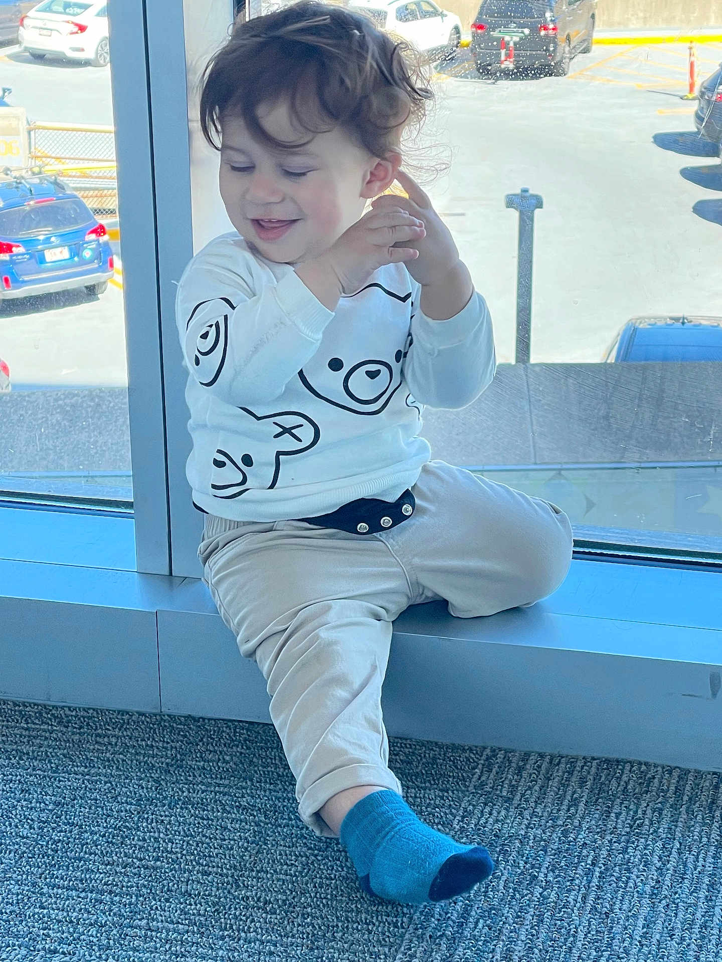 Liam a rejoint le concours — aidez-le/la à gagner de superbes lots ! toddler, child, curly_hair, sitting, window, car, parking_lot, blue_sock, beige_pants, white_sweatshirt, bear_design, happy, indoor, daylight, casual_clothing, smiling, young_child, floor_carpet, real_photo, natural_light