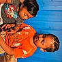 child, children, boy, toddler, sitting, blue_wall, orange_shirt, graphic_tshirt, short_hair, indoors, wooden_wall, playing, casual_clothing, brown_skin, brother_and_sister, relaxed, young, portrait, family, cute