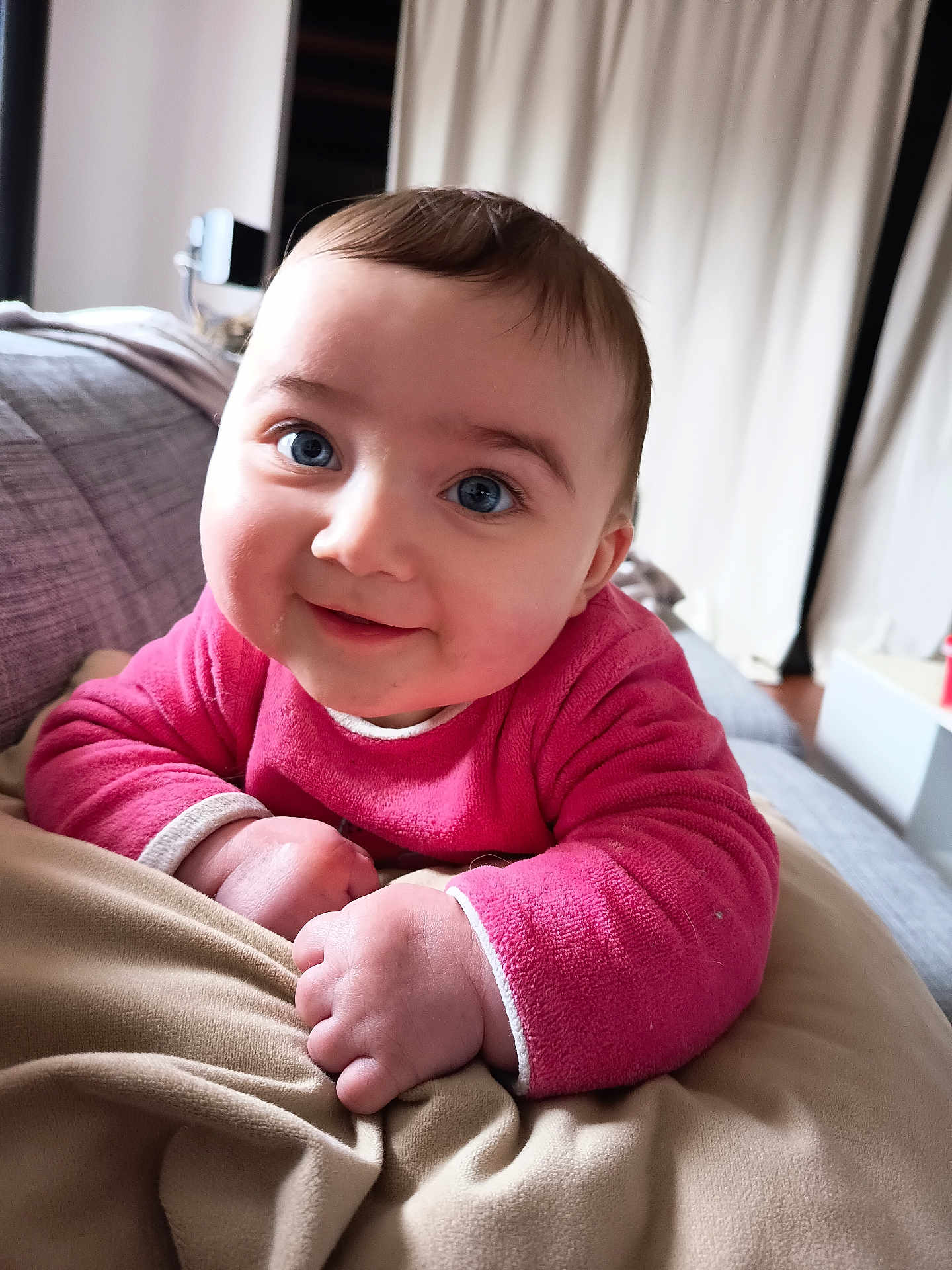 Nina a rejoint le concours — aidez-le/la à gagner de superbes lots ! baby, infant, child, portrait, close_up, blue_eyes, pink_clothing, smile, hands, cheeks, couch, blanket, indoor, curtains, face, hair, soft_texture, cozy, eye_contact, sitting