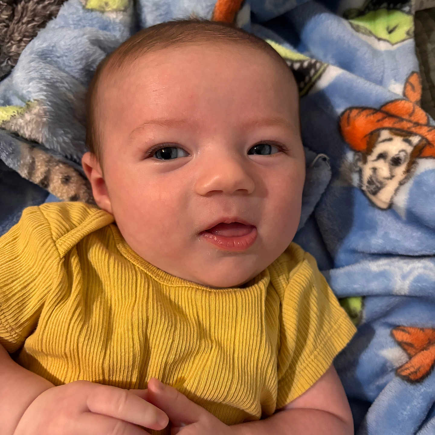 Kacee is registered to the contest to win money with this photo: baby, infant, yellow_shirt, hands, face, eyes, blanket, cartoon, soft_texture, cute, lying_down, child, skin, expression, portrait, closeup, clothing, comfort, indoors, person