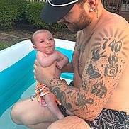 Kacee joined the competition — help win amazing prizes! baby, man, tattoos, pool, inflatable_pool, outdoor, summer, water, child, cap, swim_trunks, grass, fence, skin, holding, parent, family, cute, happy, nature