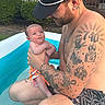 baby, man, tattoos, pool, inflatable_pool, outdoor, summer, water, child, cap, swim_trunks, grass, fence, skin, holding, parent, family, cute, happy, nature