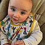 baby, infant, child, close_up, face, eyes, hands, bib, pajamas, dinosaur_print, colorful, soft_fabric, cushion, seated, indoor, cute, portrait, rosy_cheeks, curious, cozy