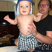 Reid is registered to the contest to win money with this photo: baby, child, woman, glasses, lip_piercing, cap, checkered_shorts, indoor, bedroom, furniture, television, cabinet, box, holding, smiling, person, skin, tattoo, happy, casual