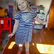 Karmah Phipps joined the competition — help win amazing prizes! child, girl, striped_dress, heart, smile, playful, indoor, sunlight, wooden_floor, red_chair, dresser, toys, socks, sneakers, blonde_hair, blue_eyes, room, furniture, casual_clothing, happy