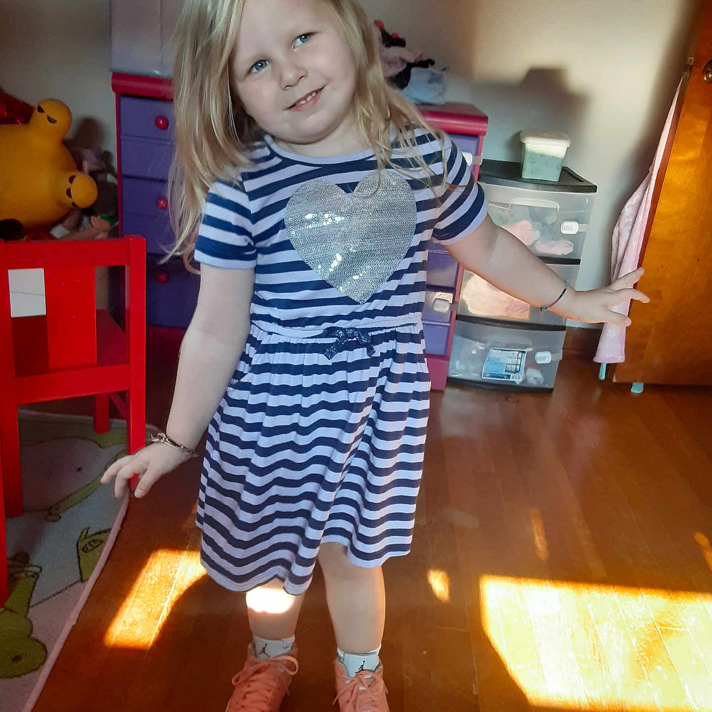 Karmah Phipps joined the competition — help win amazing prizes! child, girl, striped_dress, heart, smile, playful, indoor, sunlight, wooden_floor, red_chair, dresser, toys, socks, sneakers, blonde_hair, blue_eyes, room, furniture, casual_clothing, happy