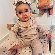 Esmée participe au concours pour gagner de l'argent avec cette photo : toddler, child, baby, pink_bows, beige_outfit, white_socks, blanket, patterned_blanket, indoor, cute, curious_eyes, soft_lighting, plush_pillow, backpack, wall, floor, sitting, hand, face, portrait