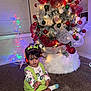 toddler, child, christmas_tree, holiday_decor, ornaments, lights, green_outfit, hair_bows, carpet, indoor, festive, decoration, smiling, holiday, seasonal, home, celebration, colorful, star_lights, winter