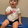 toddler, child, sitting, white_tshirt, black_shorts, bow, hair_accessory, blue_bench, curious_expression, indoor, colorful_shoes, pink, white_socks, short_hair, person, young_child, casual_clothing, looking_away, small_hands, bare_legs
