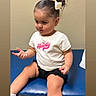 Bella is registered to the contest to win money with this photo: toddler, child, girl, bow, hair, white_shirt, black_shorts, blue_bench, indoor, portrait, cute, sitting, expression, young_child, casual_clothing, shorts, smile, skin, person, small_child