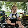 Israel is registered to the contest to win money with this photo: baby, child, birthday, crown, party, table, outdoor, grass, picnic, food, snacks, suspenders, bow_tie, shirt, decorations, park, trees, sitting, expression, celebration