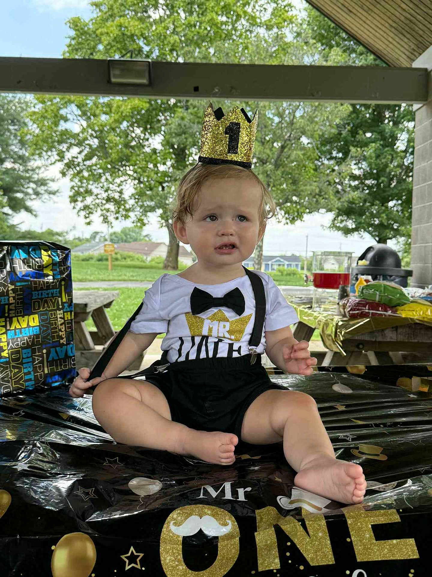 Israel is registered to the contest to win money with this photo: baby, child, birthday, crown, party, table, outdoor, grass, picnic, food, snacks, suspenders, bow_tie, shirt, decorations, park, trees, sitting, expression, celebration