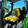 Hardin is registered to the contest to win money with this photo: baby, child, tractor, yellow_seat, green_cap, john_deere, vehicle_interior, window, trees, sky, steering_wheel, control_lever, plastic_cover, seat, clothing, person, sunlight, outdoor, portrait, serious_face