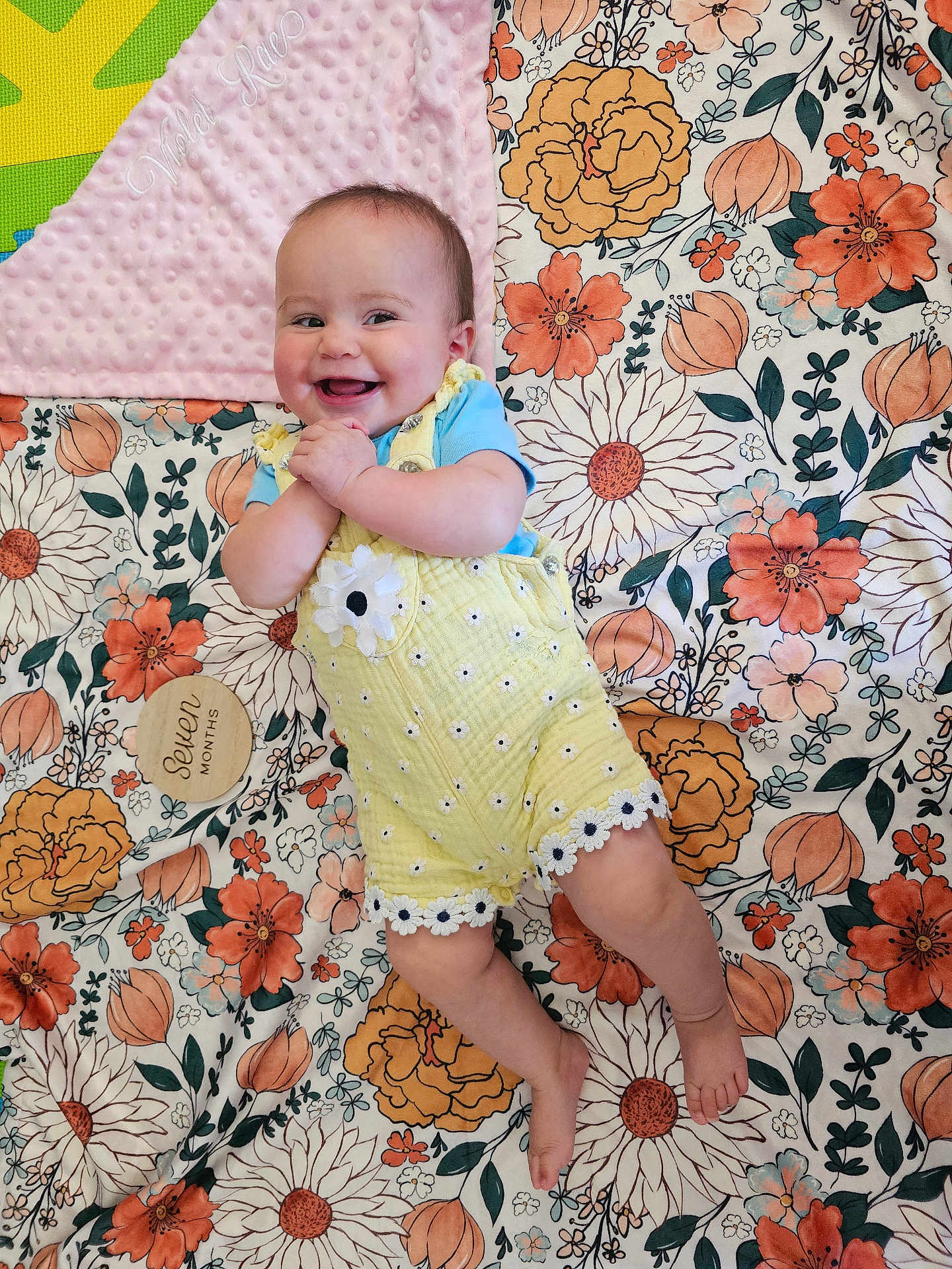 Violet joined the competition — help win amazing prizes! baby, smiling, yellow_outfit, blue_shirt, floral_blanket, pink_blanket, wooden_milestone, seven_months, happy, infant, clapping, barefoot, lying_down, cute, child, patterned_blanket, flower_pattern, indoors, portrait, young_baby