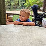 child, toddler, smile, table, outdoor, greenery, tree, stroller, chair, playful, face, person, nature, park, daylight, expression, casual, relaxing, cute, curly_hair