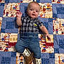 baby, child, cowboy_boots, plaid_shirt, jeans, quilt, cartoon_pattern, lying_down, smiling, cute, indoor, portrait, infant, clothing, footwear, hand_raised, colorful, fabric, person, happy
