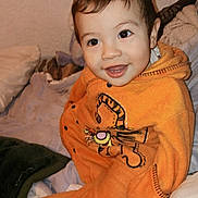 Samuel is registered to the contest to win money with this photo: toddler, child, smiling, orange_clothing, cartoon_character, blanket, pillow, indoor, happy, face, person, sitting, cute, cozy, soft_fabric, warm, home, infant, clothing, portrait