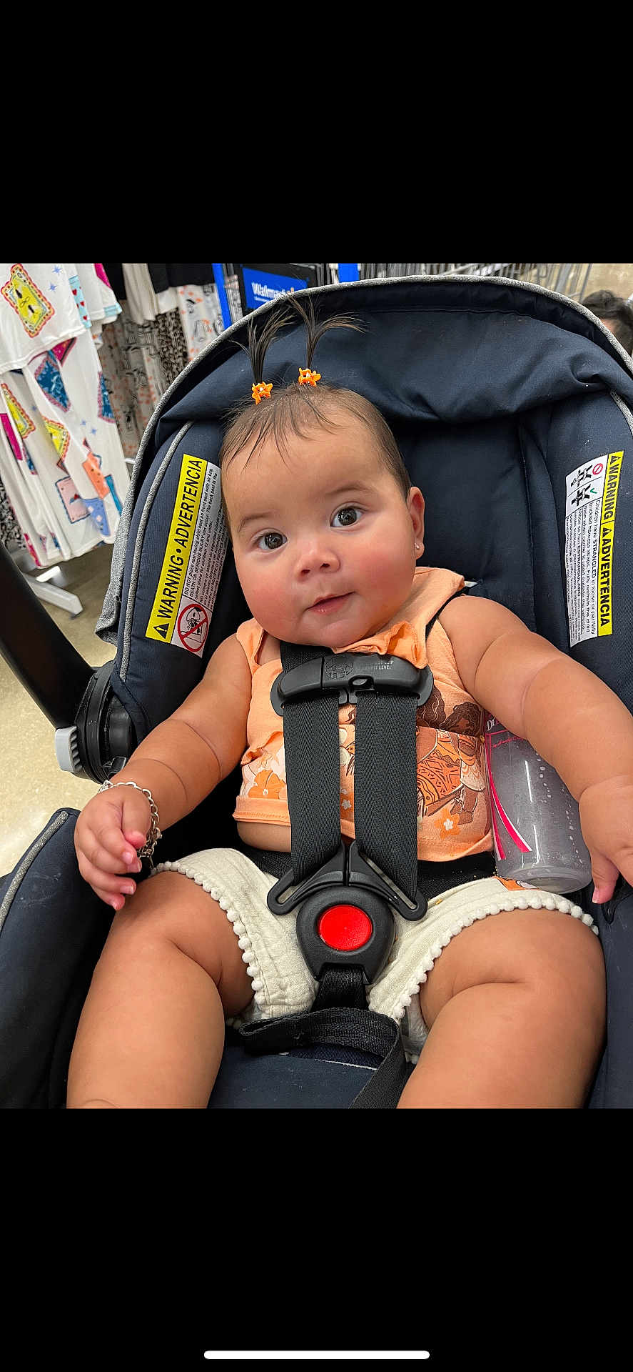 Eliana joined the competition — help win amazing prizes! baby, child, car_seat, infant, cute, smiling, hair_clips, orange, peach_top, cream_shorts, bottle, bracelet, store, clothing, shopping, curious, wide_eyes, sitting, safety_harness, indoor