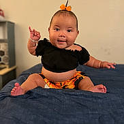 Eliana is registered to the contest to win money with this photo: baby, child, smiling, sitting, bed, blue_bedspread, black_top, orange_shorts, orange_bow, bracelet, hand_gesture, indoor, cute, happy, portrait, person, infant, skin, face, expression
