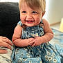 Sydney is registered to the contest to win money with this photo: baby, child, smiling, denim_dress, floral_pattern, sitting, indoors, bed, blanket, happy, face, hands, cute, expression, person, skin, blue_eyes, short_hair, infant, comfort