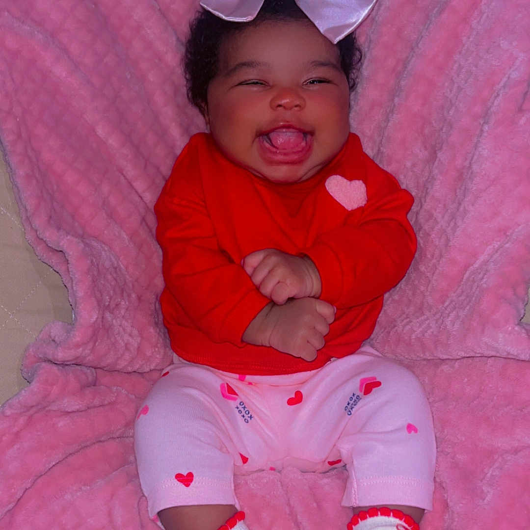 Kimaya joined the competition — help win amazing prizes! baby, bow, cheerful, child, cozy, cute, footwear, happy, heart_pattern, indoors, infant, person, pink_blanket, portrait, red_clothing, sitting, smiling, sock, soft_texture, warm_colors