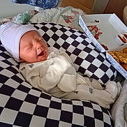 Mack is registered to the contest to win money with this photo: newborn, baby, infant, hat, onesie, checkered_cushion, food_box, fries, sauce, bedroom, quilt, blanket, sleeping, yawning, cozy, indoor, cute, person, resting, soft_lighting