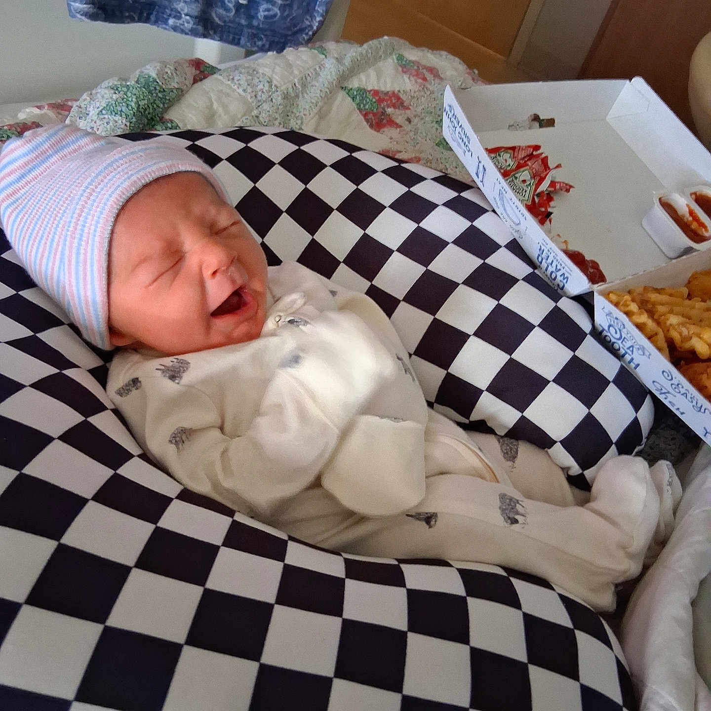 Mack is registered to the contest to win money with this photo: newborn, baby, infant, hat, onesie, checkered_cushion, food_box, fries, sauce, bedroom, quilt, blanket, sleeping, yawning, cozy, indoor, cute, person, resting, soft_lighting