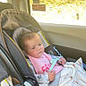 baby, infant, child, car_seat, vehicle, travel, passenger, sitting, face, portrait, serious_expression, pink_clothing, blanket, toy, teether, sunlight, window, motion_blur, indoor, road_trip