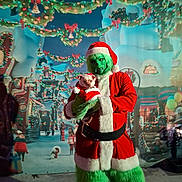 Simba is registered to the contest to win money with this photo: person, costume, grinch, dog, pet, holiday, christmas, santa_outfit, festive, lights, decorations, background, celebration, winter, green_fur, red_clothing, small_dog, holding, outdoor, night