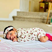 Anshika is registered to the contest to win money with this photo: baby, infant, lying_down, blanket, headband, floral_outfit, pink_socks, indoors, carpet, face, person, cute, child, young_child, home, soft_lighting, portrait, looking, resting, cozy
