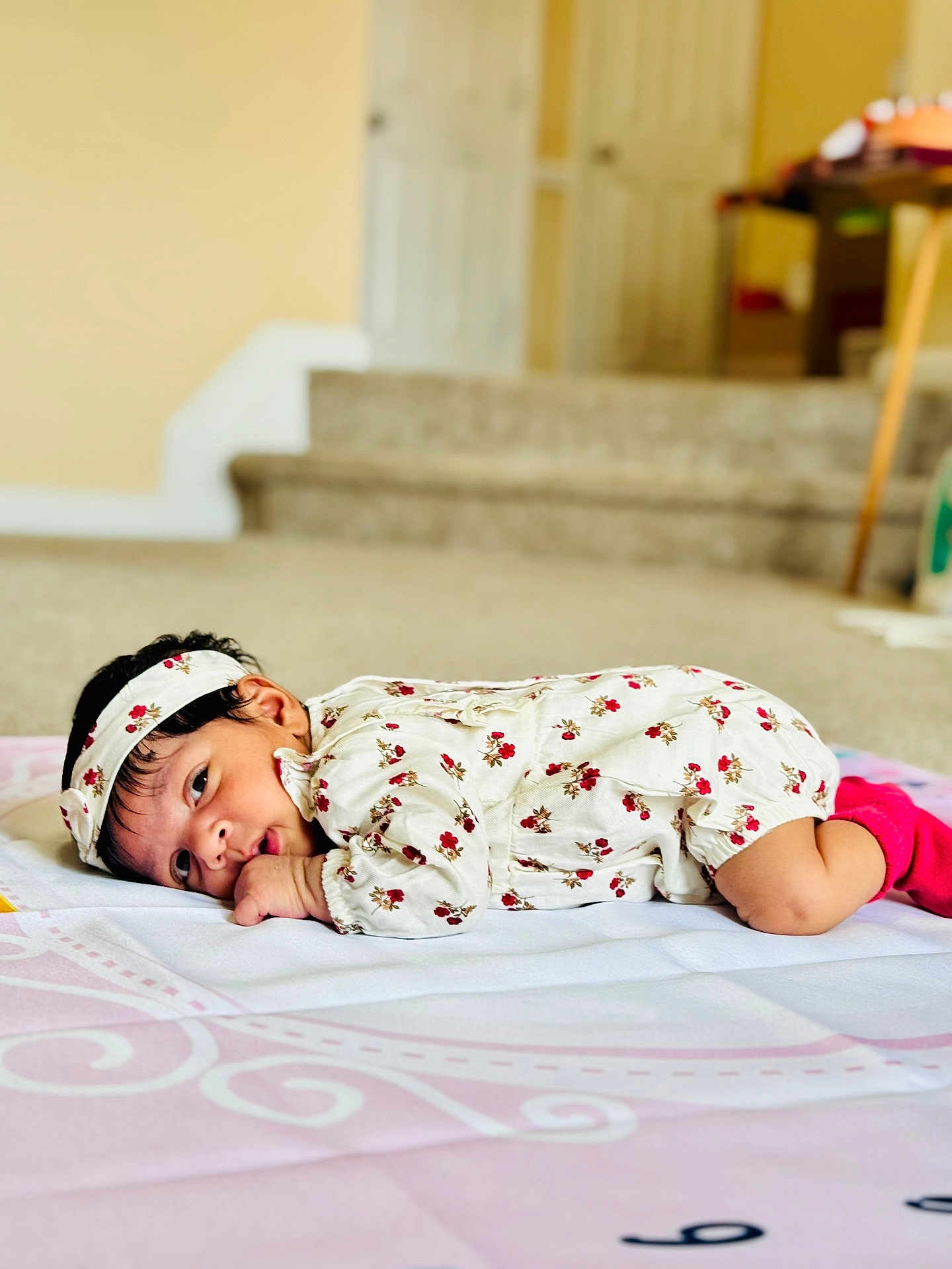 Anshika is registered to the contest to win money with this photo: baby, infant, lying_down, blanket, headband, floral_outfit, pink_socks, indoors, carpet, face, person, cute, child, young_child, home, soft_lighting, portrait, looking, resting, cozy