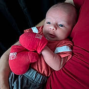 Elias is registered to the contest to win money with this photo: baby, infant, mittens, red_clothing, grey_pants, adult_arm, cradling, close_up, indoor, portrait, newborn, cute, human, child, soft_light, cozy, expression, holding, person, warm