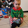 celebration, child, christmas_decor, christmas_tree, cute, decorations, festive, floor_tiles, gift, green_clothing, holiday, holiday_outfit, holiday_spirit, indoors, presents, red_and_white_stripes, sitting, toddler, wrapped_gift, young_child