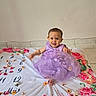 baby, child, smiling, purple_dress, flower, blanket, indoor, tiled_floor, cute, portrait, happy, sitting, decoration, floral_pattern, infant, person, celebration, bright_colors, adorable, toddler